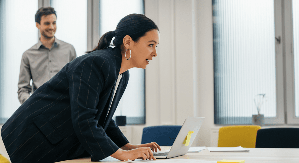 A woman in a pinstripe blazer works on a laptop while a man observes in a modern, brightly lit office setting.